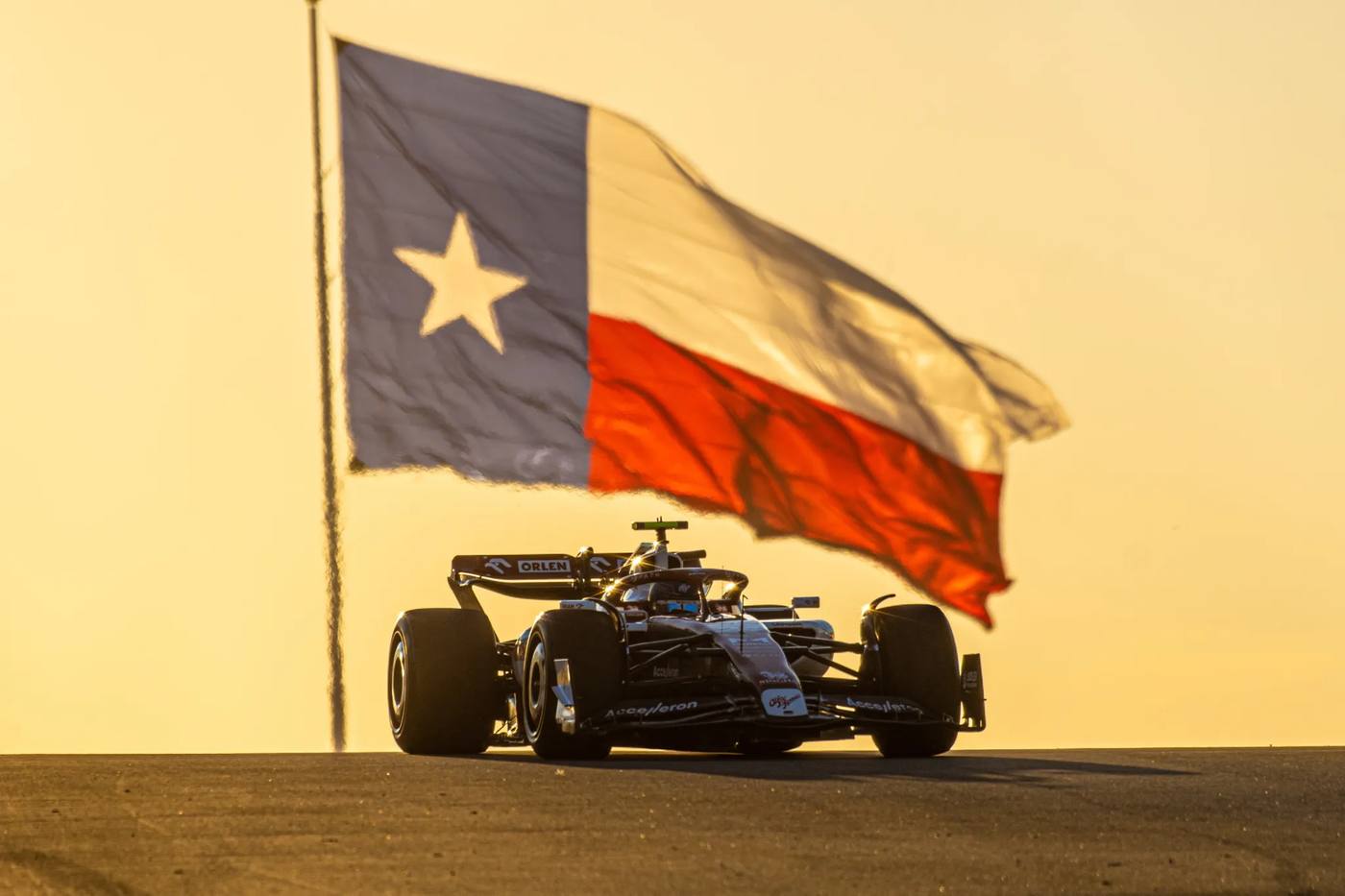 A Formula 1 car crests a hill at Circuit of the Americas at golden hour, the Texas flag flying behind it