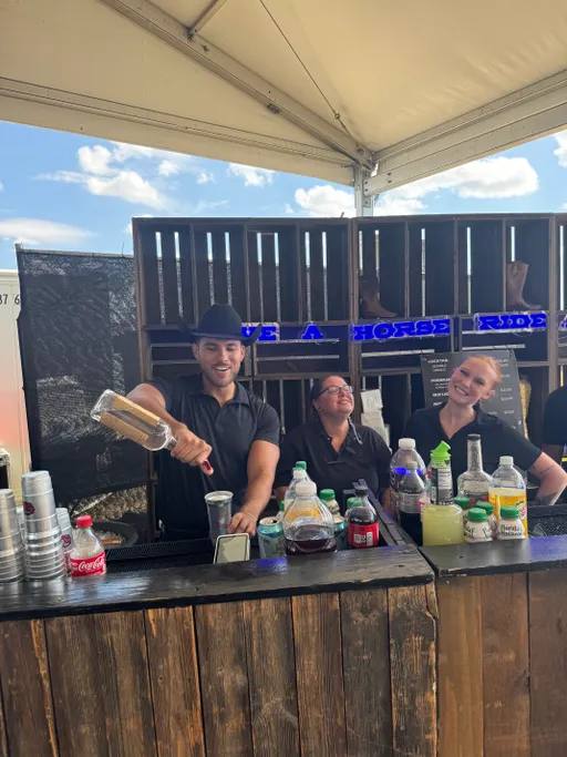 Three Undergrads bartenders in black behind a wooden bar under a tent at COTA
